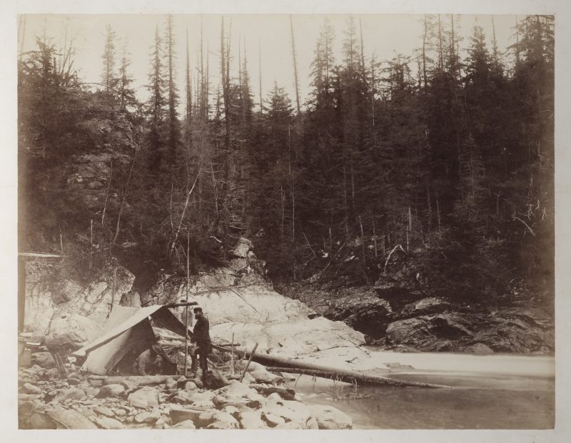 A man stands on a rocky beach in front of canvas tent.