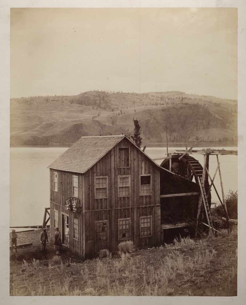 A wooden mill with a large water wheel attached to the side sits on a rocky beach.